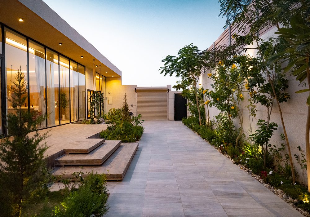 Modern house with glass walls and doors, a walkway to the front door, plants, trees, stones, and lights
