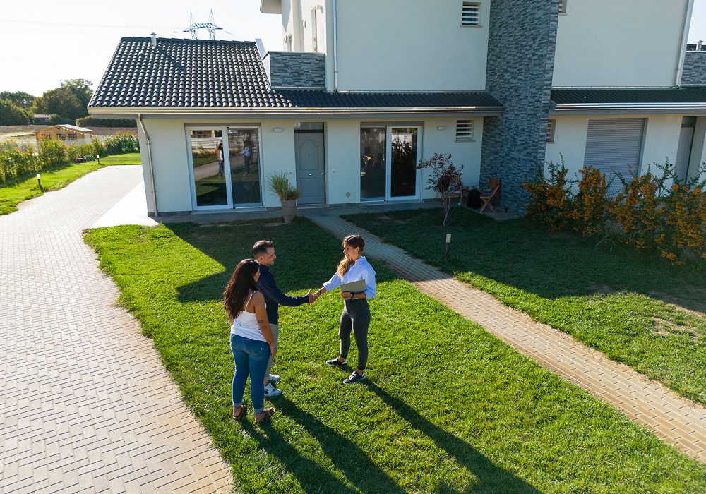 Couple shaking hands in front of a house surrounded by lush green grass, plants, and trees