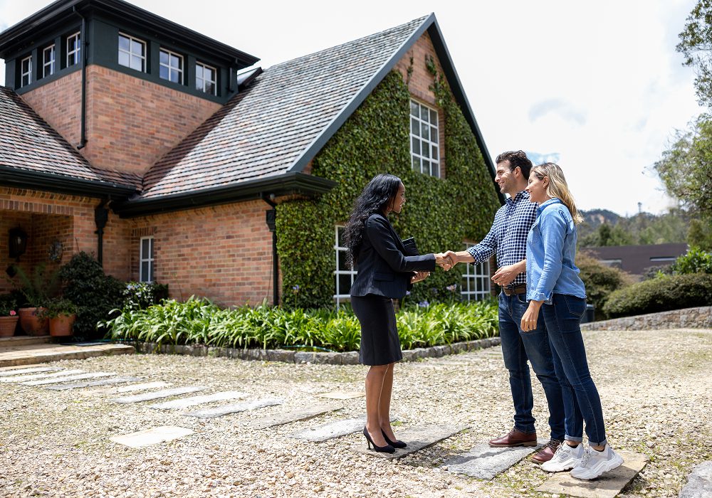 Couple shaking hands in front of a house surrounded by lush greenery, with trees, plants, and flowers in the background