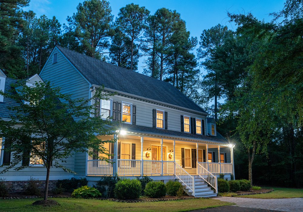Large white house with a porch and a driveway surrounded by trees, plants, and grass