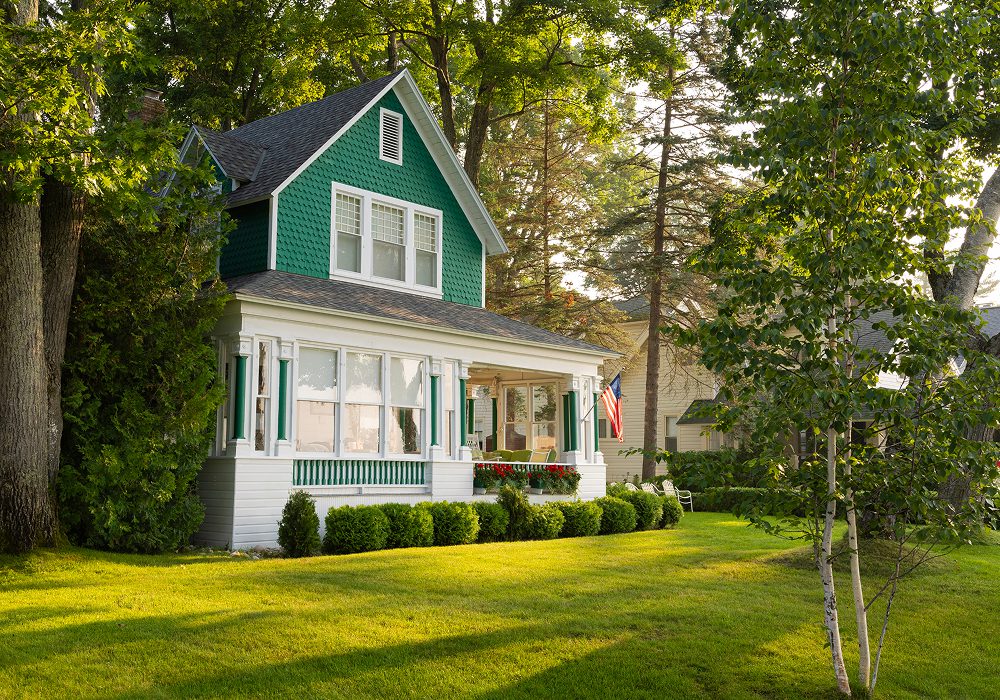 Green house with white trim and a large lawn surrounded by trees, plants, and grass