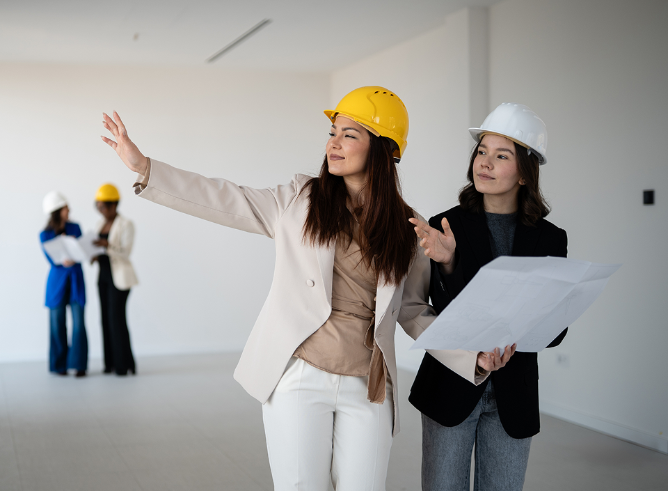 Two women in hard hats standing in an empty room, looking at blueprints